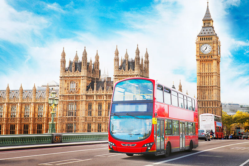 A red London double-decker bus driving across Westminster Bridge, with the Houses of Parliament in the background.